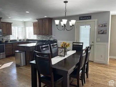 Kitchen featuring dark-toned cabinetry, a stone-patterned backsplash, wood-finish flooring, a tiered chandelier, and wainscoting detailing
