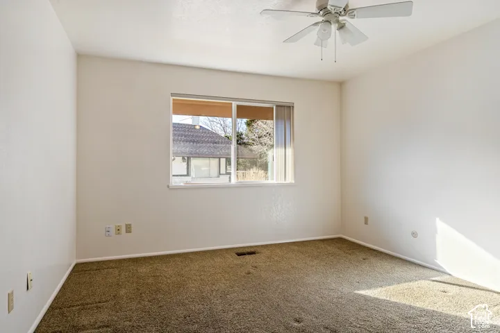 Neutral-toned interior space featuring carpeted flooring, a ceiling fan with integrated lighting, and a single-pane sliding window with vertical blinds