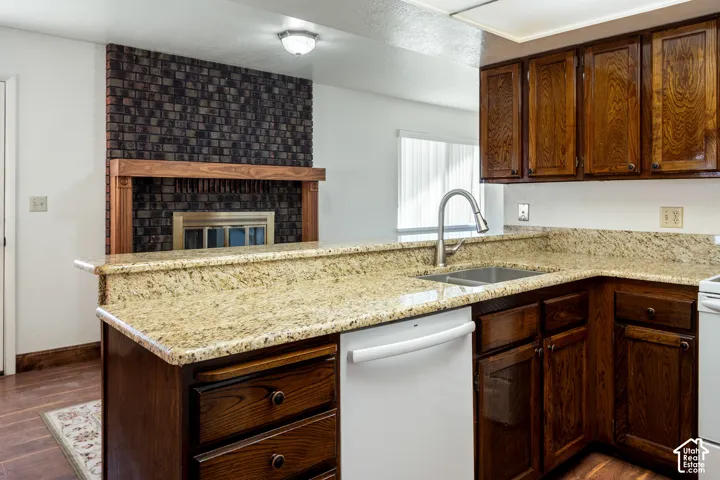 Kitchen featuring stone-finish countertops, dark wood cabinetry, a stainless steel dual-basin undermount sink with a gooseneck faucet, and an exposed brick fireplace with a wood mantel