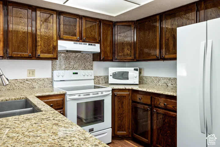 Kitchen featuring stained wood cabinetry, stone-finish countertops, stone-finish backsplash, built-in white range with overhead vent hood, and a dual-basin undermount sink