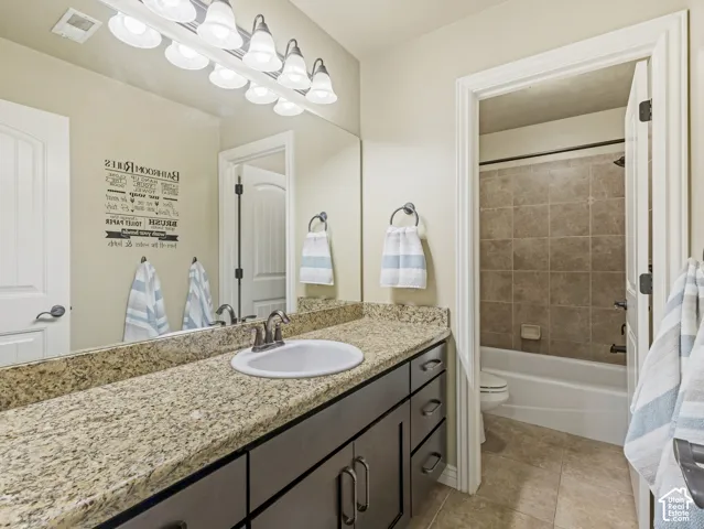 Bathroom featuring a stone-finish countertop with an undermount sink, dark-toned cabinetry, multi-bulb vanity lighting, and a bathtub with full-height tile surround