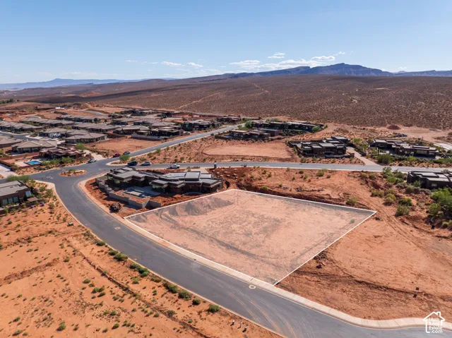 Expansive undeveloped lot with paved road frontage, bordered by established residences and desert landscapes, and distant mountains
