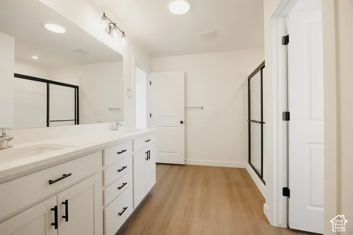 Dual-sink vanity with a solid-surface countertop and matte black hardware, wood-finish flooring, full-width mirror, and a walk-in shower with a black-framed glass enclosure