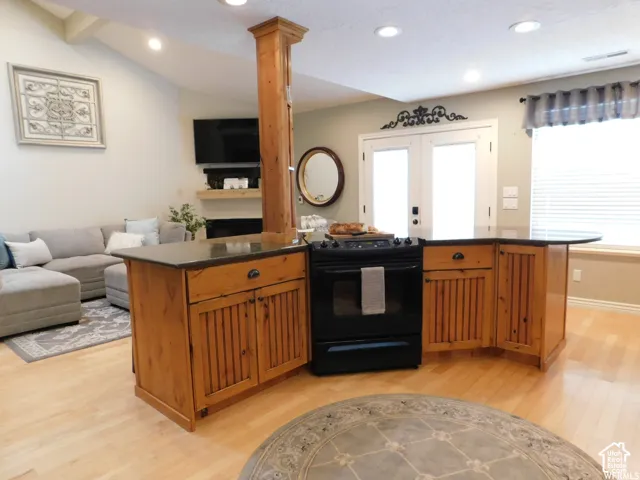 Kitchen island featuring wood-finish cabinetry, a dark stone countertop, and a built-in range, complemented by light wood-finish flooring, vaulted ceilings, and double glass doors