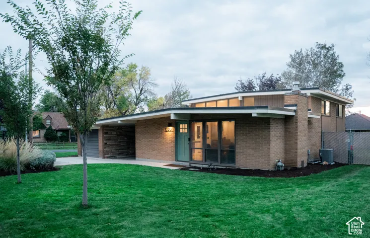 Mid-century modern exterior featuring a low-pitch roofline, brick masonry facade, integrated carport with stone-finish accent wall, and expansive floor-to-ceiling windows