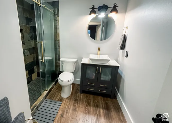 Bathroom featuring wood-finish flooring, a glass-enclosed shower with tiled walls and pebble-style floor, a vanity with a rectangular vessel sink and brass-tone faucet, and a round wall-mounted mirror