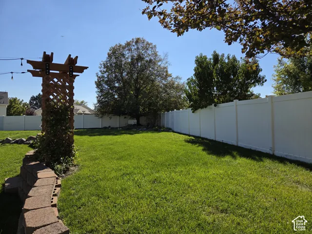 Spacious backyard featuring a white vinyl privacy fence, a natural stone retaining wall, a wooden lattice pergola, and mature trees