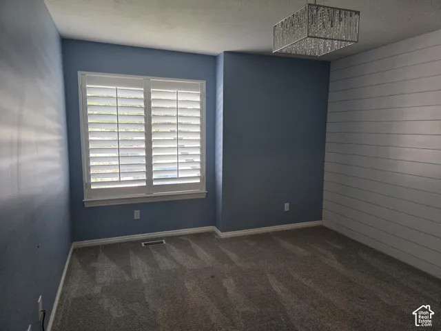 Room featuring a white shiplap accent wall, neutral carpeting, and a rectangular tiered chandelier with glass elements, complemented by interior window shutters