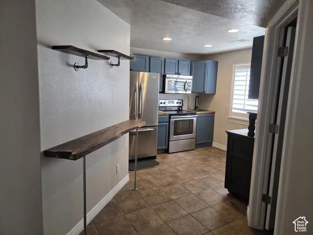 Kitchen featuring blue-toned cabinetry, stainless steel appliances, custom live-edge wood shelving, a live-edge wood counter, and tiled flooring