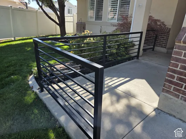 Concrete patio featuring a matte black horizontal metal railing, brick masonry accent, and landscaped garden beds
