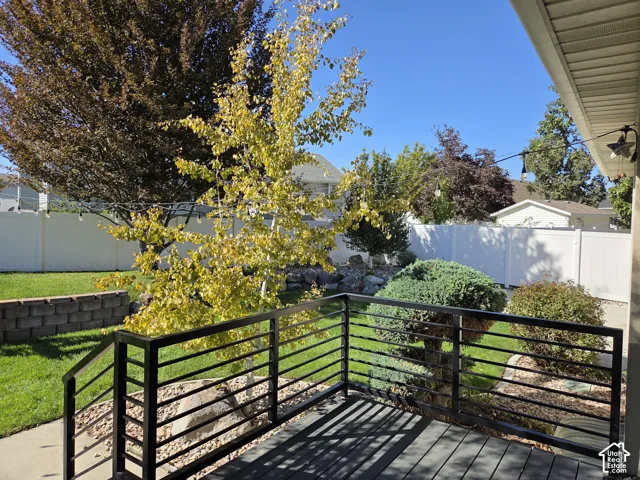 Backyard featuring a raised deck with black metal railing, stone-block retaining wall, manicured lawn, and perimeter vinyl fencing