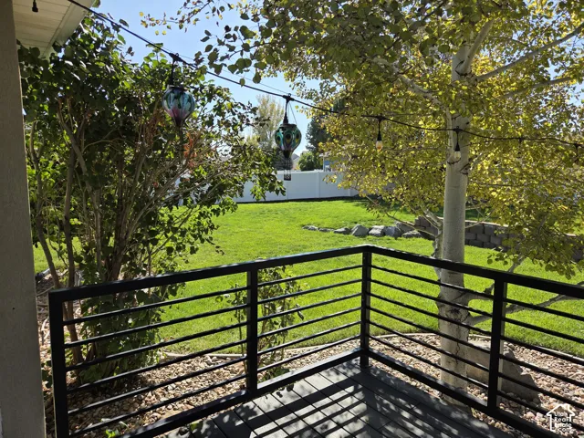 Covered deck featuring black metal railings, overlooking a backyard with a manicured lawn, mature trees, and a decorative stone retaining wall