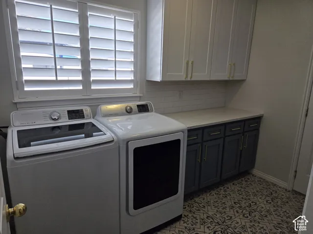 Laundry space featuring patterned tile flooring, charcoal base cabinetry with gold-tone hardware, white upper wall-mounted cabinetry, a stone-finish countertop, and window shutters