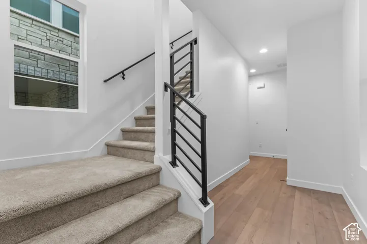 Staircase featuring plush carpeted steps, matte black metal railings, wood-finish flooring, recessed lighting, and a window with exterior stone masonry detail