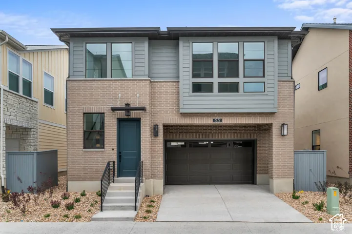 Two-story exterior featuring tan brick masonry, grey horizontal siding, a multi-pane grid window design, an attached garage, and a concrete stoop with metal railing