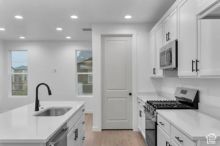 Kitchen featuring white shaker-style cabinetry, quartz countertops, an island with an undermount sink and matte black faucet, stainless steel appliances, and wood-finish flooring