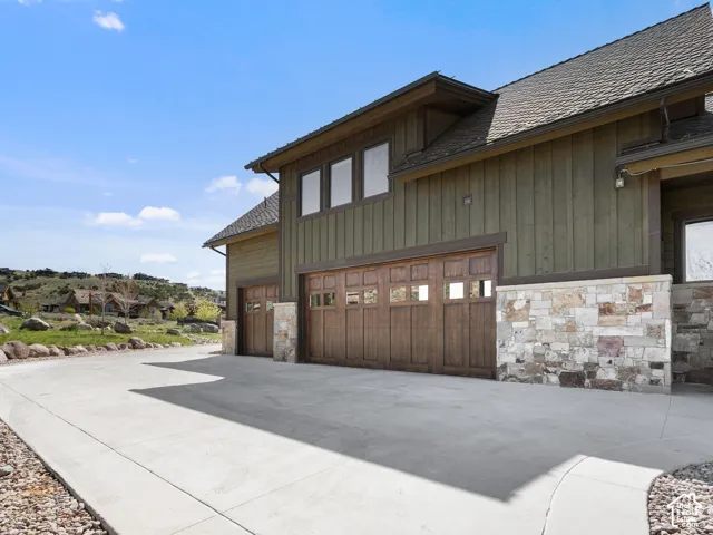 Exterior featuring vertical board-and-batten siding, stone masonry wainscoting, expansive concrete driveway, and custom multi-pane wood garage doors