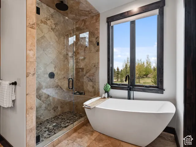 Walk-in shower featuring large-format tile surround, pebble flooring, and a frameless glass enclosure, paired with a freestanding soaking tub, matte black plumbing fixtures, and dark-stained wood window trim