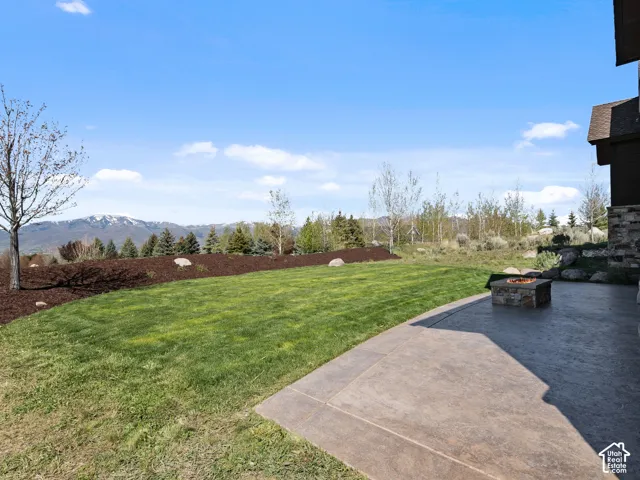 Stamped concrete patio featuring a built-in stone fire pit, grass lawn, mulched garden beds, and mountain scenery