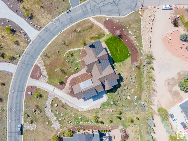Aerial view of a residential property featuring a multi-gabled shingle roof, integrated metal roofing accents, a paved concrete driveway, a manicured grass lawn, and xeriscaped grounds with natural rock formations