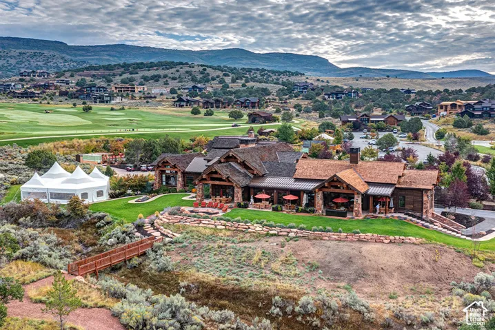 Rustic estate exterior featuring stone and wood-shake siding, covered patios, a manicured lawn, and an arched wooden footbridge set against a mountainous horizon