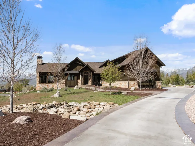 Custom residence featuring stone masonry and dark-toned siding, asphalt shingle roofing, prominent stone chimney, and an expansive concrete driveway