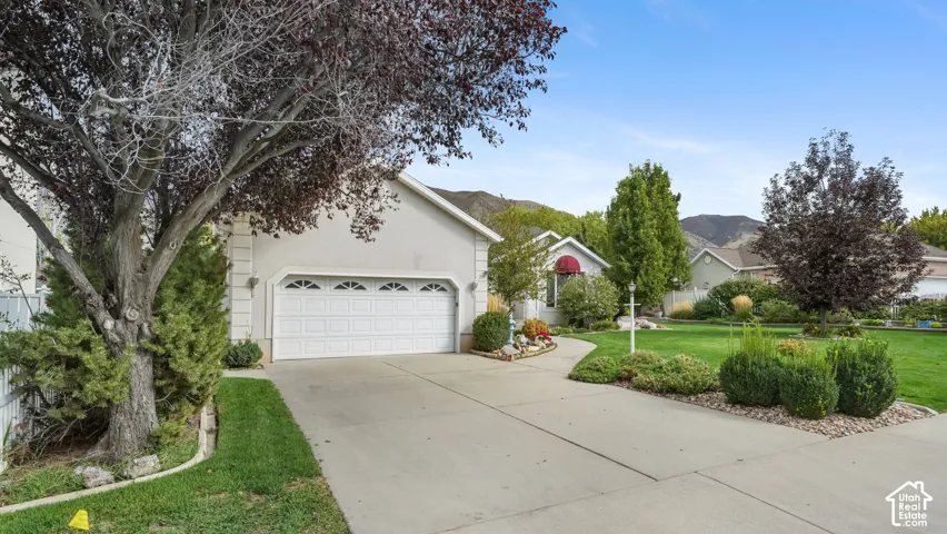 Stucco exterior featuring a two-car garage with arched windows, concrete driveway, and established landscaping with mature trees and shrubbery against a mountain backdrop