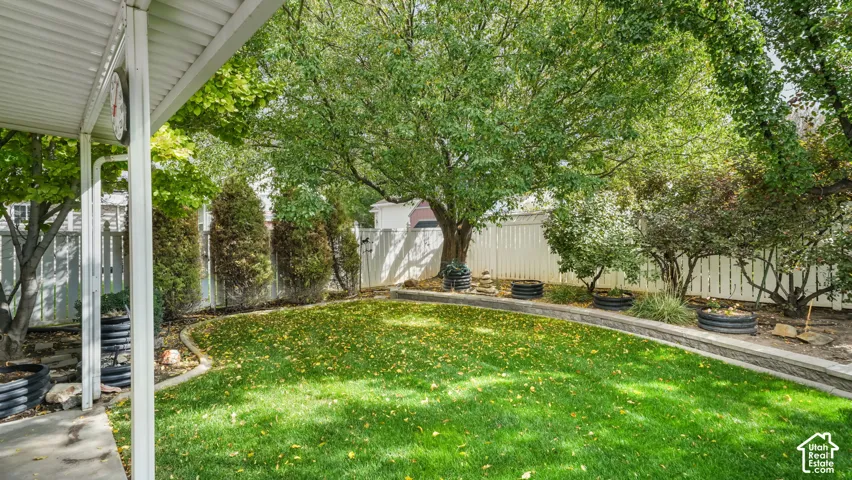 Backyard featuring a covered patio, a manicured lawn, tiered stone retaining walls, and mature landscaping enclosed by white vinyl fencing
