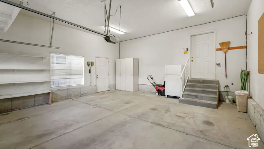 Garage space featuring concrete flooring, built-in wall shelving, enclosed storage cabinetry, a single window, and a staircase with a handrail