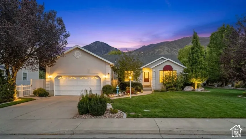 Single-story residence featuring a light-toned exterior, attached two-car garage, concrete driveway, and manicured front lawn set against a mountain backdrop