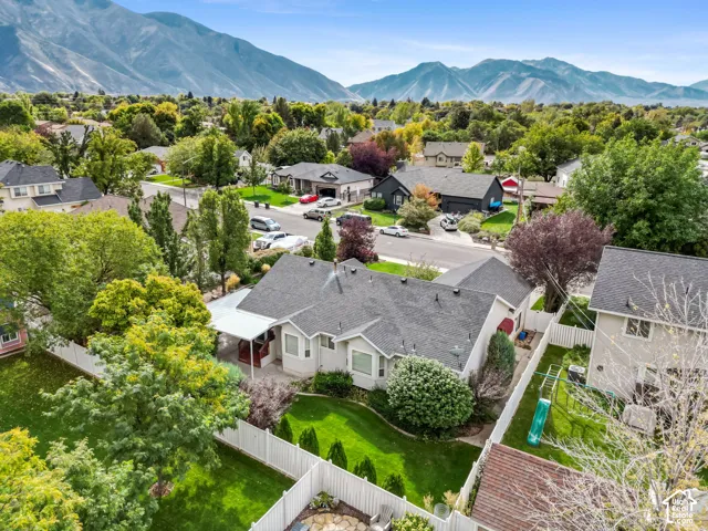 Aerial perspective featuring a residential lot with a manicured lawn, white vinyl perimeter fencing, a paved patio, and mountain scenery