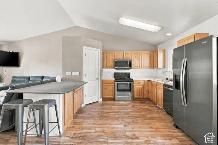 Kitchen featuring natural wood cabinetry, stainless steel appliances, wood-finish flooring, a vaulted ceiling, and a peninsula with a matte-finish countertop