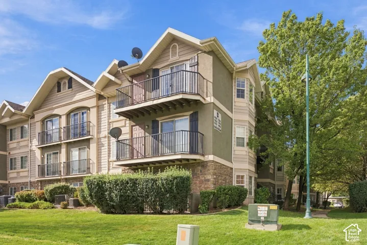 Multi-story building featuring stone veneer foundation, neutral-toned siding, private metal-railed balconies, and gabled rooflines surrounded by manicured landscaping