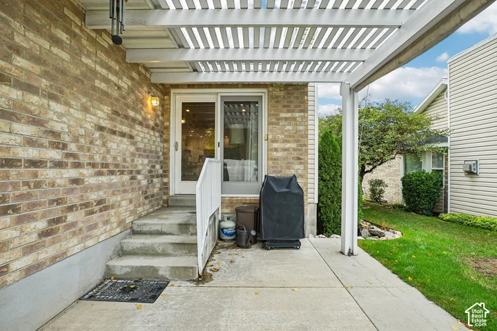 Covered patio featuring a white slatted pergola, concrete flooring, brick exterior masonry, and a glass-paneled entry door with metal railing
