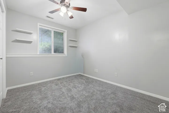 Room interior featuring carpeted flooring, a ceiling fan with integrated lighting, wall-mounted floating shelving, and a double-hung window with horizontal blinds
