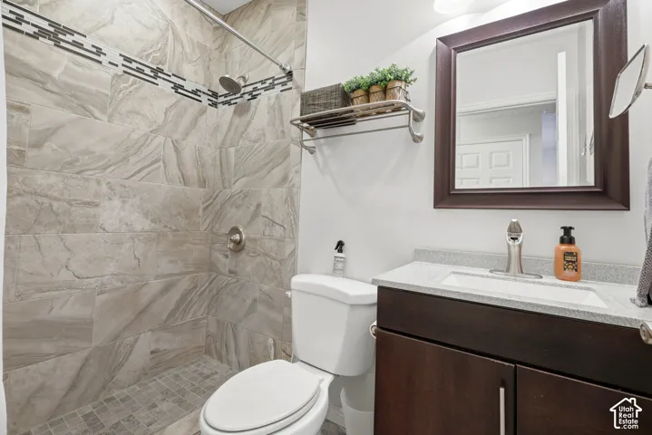 Bathroom on the first level featuring a tile shower surround with a decorative mosaic accent border, a dark wood-finish vanity with a stone-composite countertop, and a wall-mounted metal shelving unit