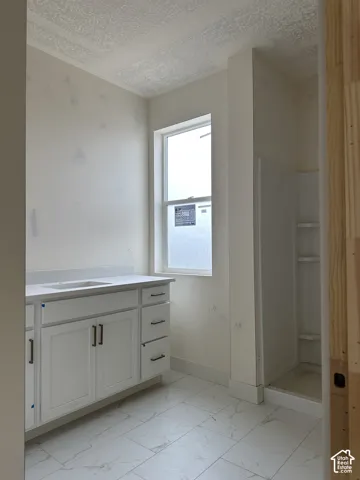 White vanity cabinet featuring rectangular basin, quartz countertop, and dark metal hardware, complemented by floor-to-ceiling tile-patterned flooring and a stand-alone shower enclosure with integrated shelving