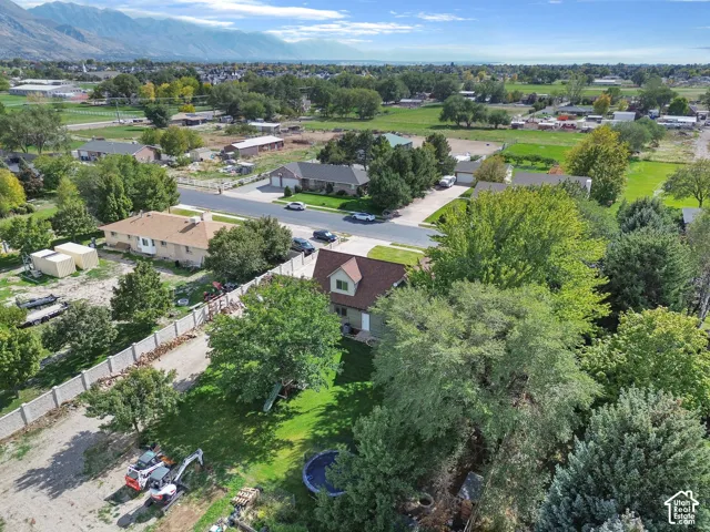 Aerial perspective showcasing a parcel with mature trees, a manicured lawn, a gravel parking area, a perimeter fence, and mountain scenery
