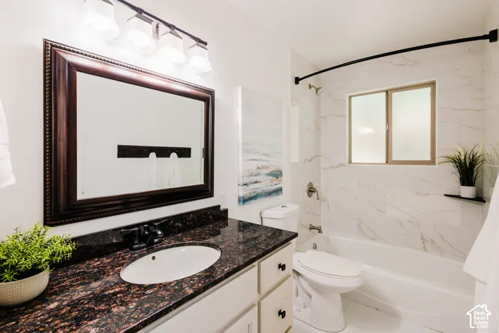 Bathroom featuring a vanity with a dark-toned countertop, white cabinetry with black hardware, a framed wall mirror, four-light vanity fixture, and a tub-shower combination with floor-to-ceiling stone-finish wall tile