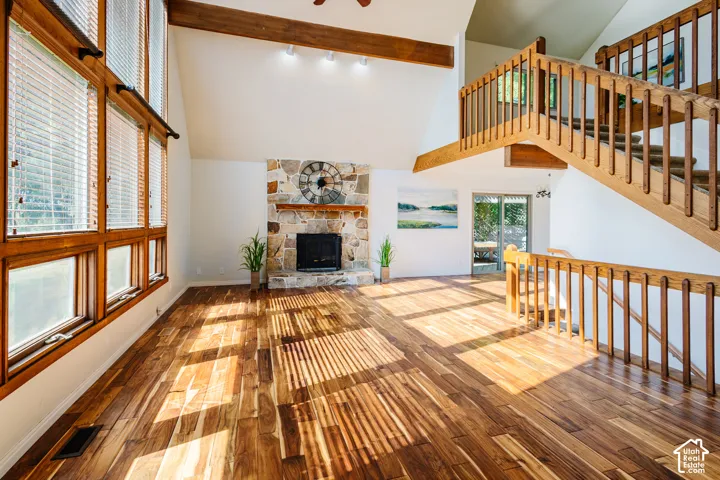 Vaulted ceiling with exposed wood beams, stone-surround fireplace, floor-to-ceiling windows, wood-finish flooring, and wood stair railing