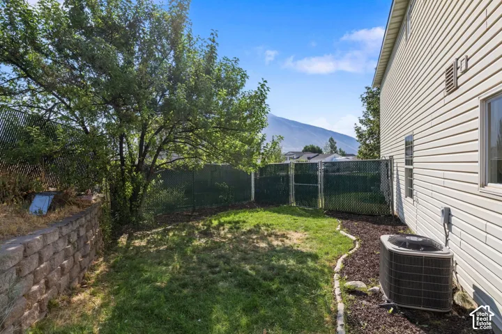 Rear yard featuring a lawn, mature trees, a segmented block retaining wall, and mountain scenery