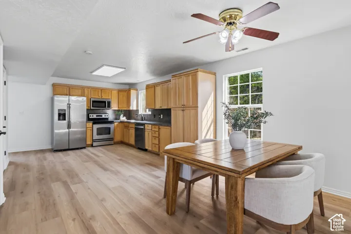 Kitchen featuring natural wood cabinetry, stainless steel appliances, dark countertops, wood-finish flooring, and a multi-light ceiling fan