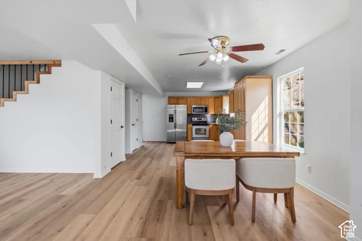 Open-concept interior featuring wood-finish flooring, natural wood cabinetry, stainless steel appliances, a ceiling fan with integrated lighting, and a staircase with metal balusters