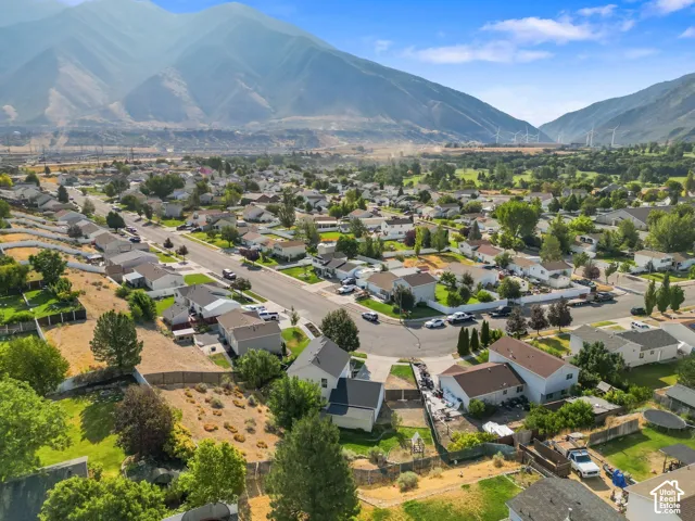 Aerial perspective of the residential property featuring mountain scenery, landscaped grounds with mature trees, paved street access, and a concrete driveway