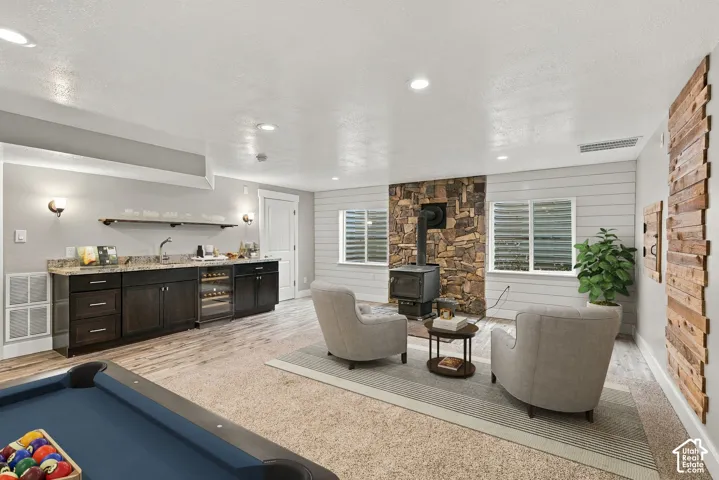 Wet bar featuring dark cabinetry, stone-finish countertops, stainless steel sink, and built-in wine refrigerator, complemented by a floor-to-ceiling natural stone fireplace surround, wood-plank accent wall, recessed lighting, and wood-finish flooring