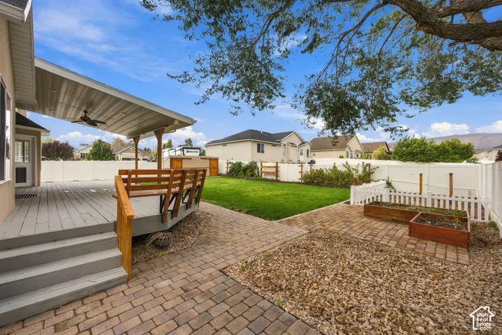 Covered wooden deck featuring a ceiling fan, stone-paver patio, grass lawn, raised timber garden beds, and privacy fencing