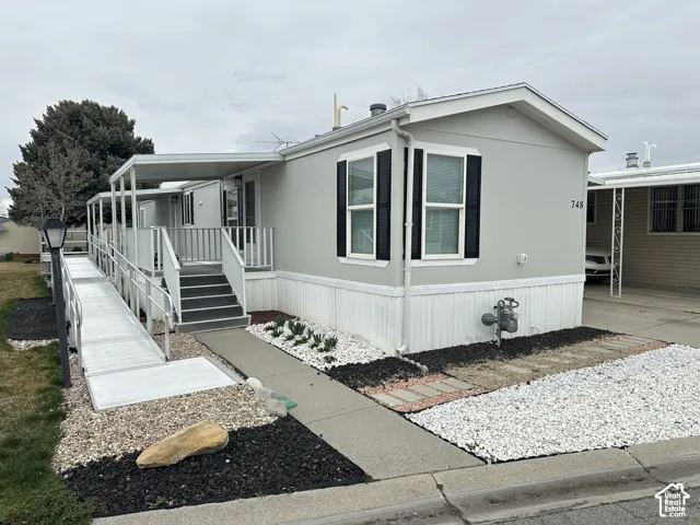 Exterior featuring light-toned siding with contrasting window shutters, a covered porch, an metal access ramp, and low-maintenance stone landscaping