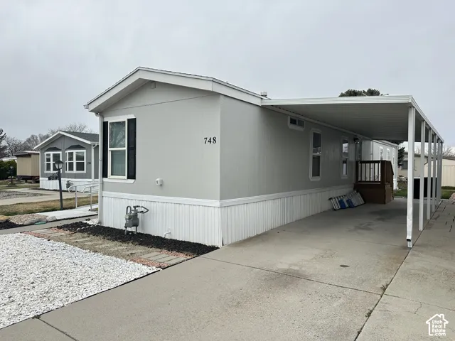 Light gray exterior siding featuring black window shutters, a concrete driveway, an attached carport, and a raised wooden entry deck