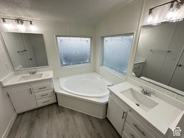 Dual white vanities with rectangular under-mount sinks, corner soaking tub, patterned privacy glass windows, and wood-finish flooring