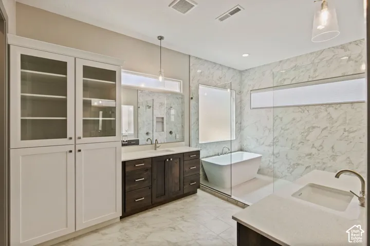 Bathroom featuring dual wooden vanities with quartz countertops, stone-finish wall cladding, a glass-enclosed shower, and a freestanding soaking tub